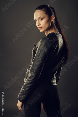 Платно Beautiful girl in black leather jacket and black leggings posing on a gray background