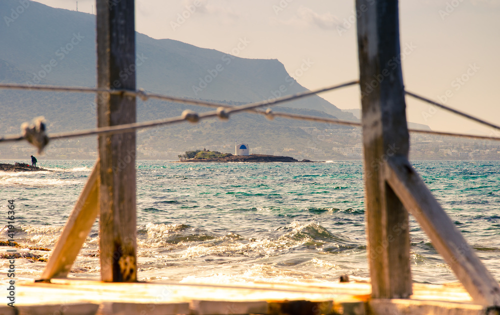 Obraz premium Typical summer image of an amazing pictorial view through a frame of a wooden pier and ropes of an old white church in a small island, Malia, Crete, Greece.