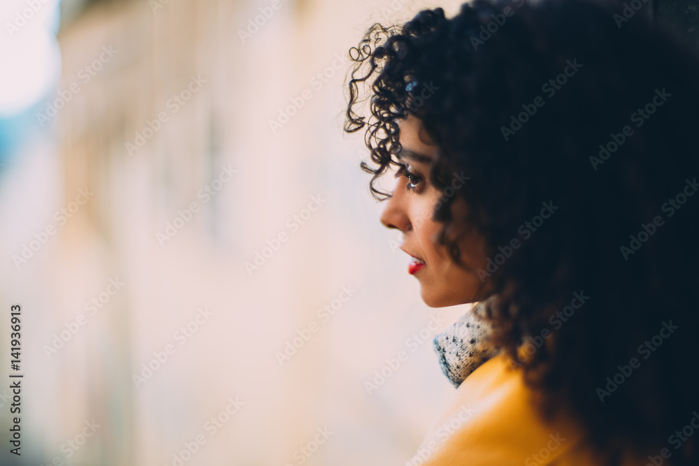 Fototapeta premium True tilt shift shooting of beautiful pensive Brazilian woman with curly hair and yellow coat looking away, blurred background with copy space for young advertising text message or promotional content