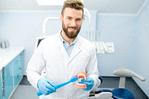 Portrait of handsome dentist in uniform with big toothbrush and artificial jaw at the dental office