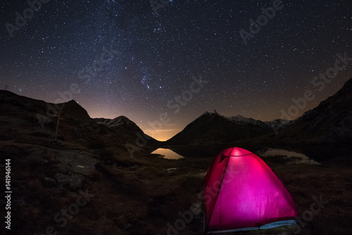 Camping under starry sky and Milky Way arc at high altitude on the italian french Alps. Glowing tent in the foreground. Adventure into the wild.