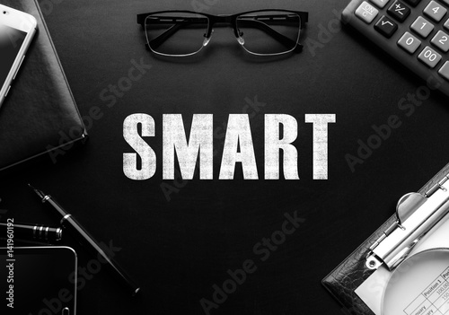 Black chalkboard with business accessories (notepad, magnifying glass, fountain pen, tablet, phone, glasses and calculator) and text SMART. Top view.