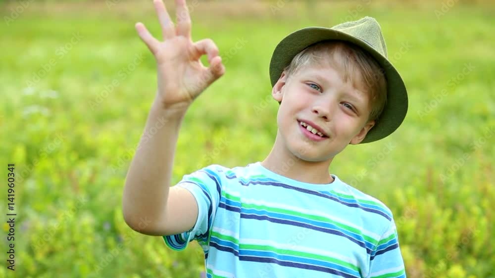 Portrait of cute funny blond 8 years old boy looking at camera smiling outside isolated on green grass background. Child shows ok sign with fingers. Real time full hd video footage. Portrait of cute funny blond 8 years old boy looking at camera smiling outside isolated on green grass background. Child shows ok sign with fingers. Real time full hd video footage.
