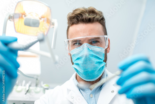 Portrait of scary dentist in mask and protective eyeglasses with dental tools during the surgery looking at camera