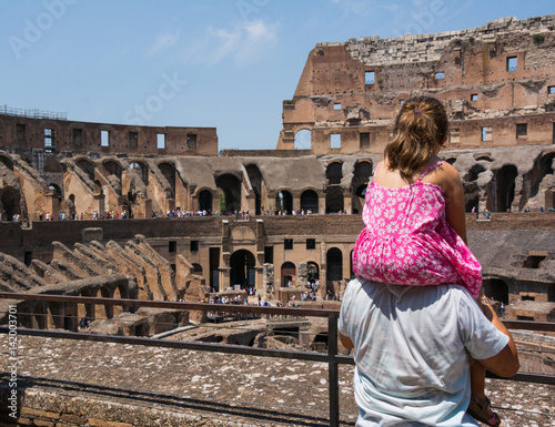 Father is holding his daughter to see the inside arena of Coliseum on a sunny hot summer day. Family observing ancient construction of Colloseo, a popular tourist destination photo