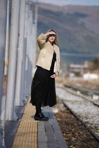 Blond young woman standing at the edge railway platform waiting wear sheep jacket and black long skirt