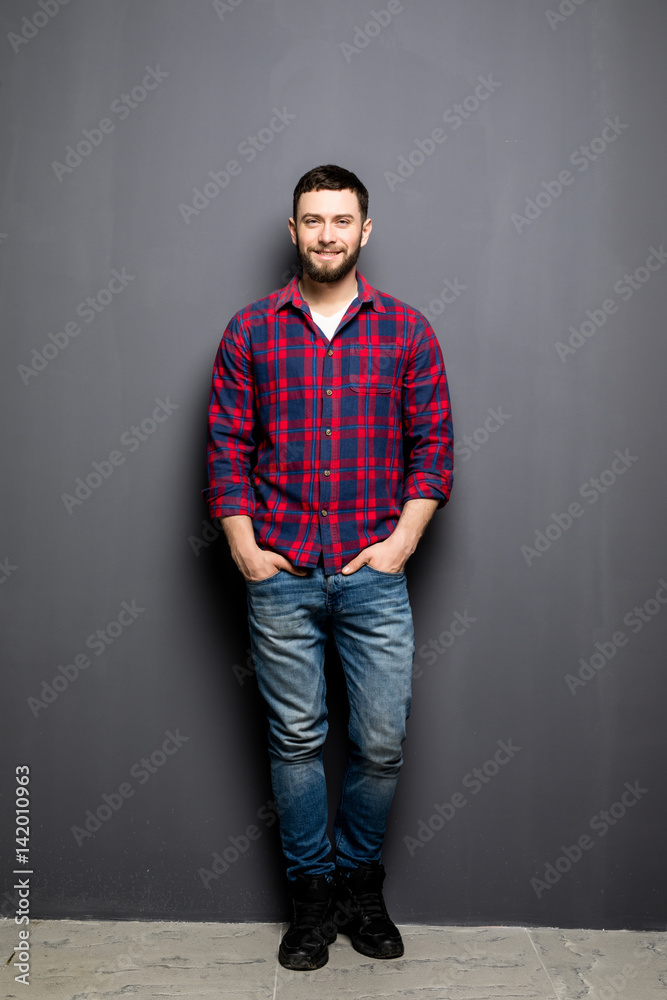 Full Portrait of handsome young man in casual shirt with hands in pockets and smiling while standing against grey background Full Portrait of handsome young man in casual shirt with hands in pockets and smiling while standing against grey background