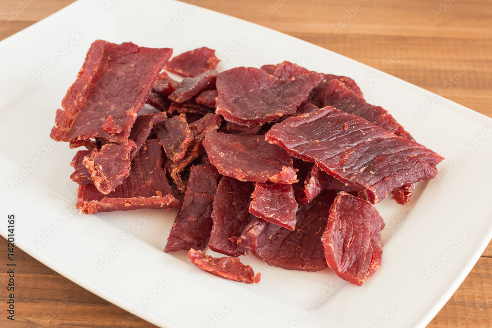 Fototapeta premium Beef jerky pieces on white dish on wooden background.