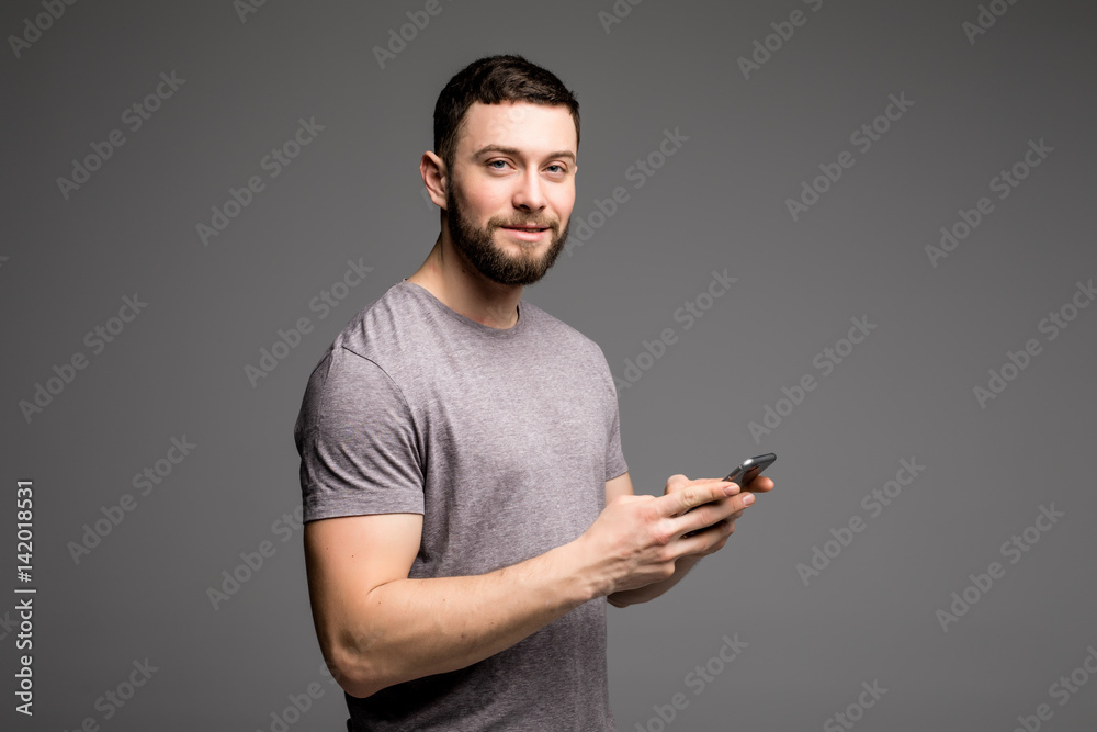Handsome young man in shirt holding mobile phone and looking at camera while standing against grey background Handsome young man in shirt holding mobile phone and looking at camera while standing against grey background