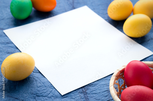 Multicolored easter eggs, basket, and a piece of paper on a textured blue background. The concept of a holiday and a happy Easter. With space for text