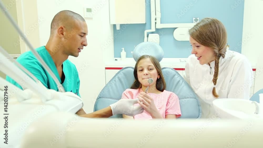 Friendly male dental surgeon and female dentist showing mouth mirror to cute little girl before examining her Friendly male dental surgeon and female dentist showing mouth mirror to cute little girl before examining her