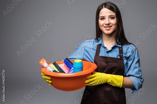 Young woman holding cleaning tools and products in bucket, isolated on grey