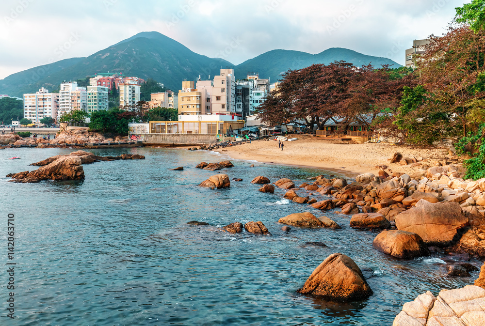 Naklejka premium Skalisty brzeg morza i mała piaszczysta plaża Zatoki Stanleya w Hongkongu. Piękny krajobraz z wodą, górami, skałami i budynkami