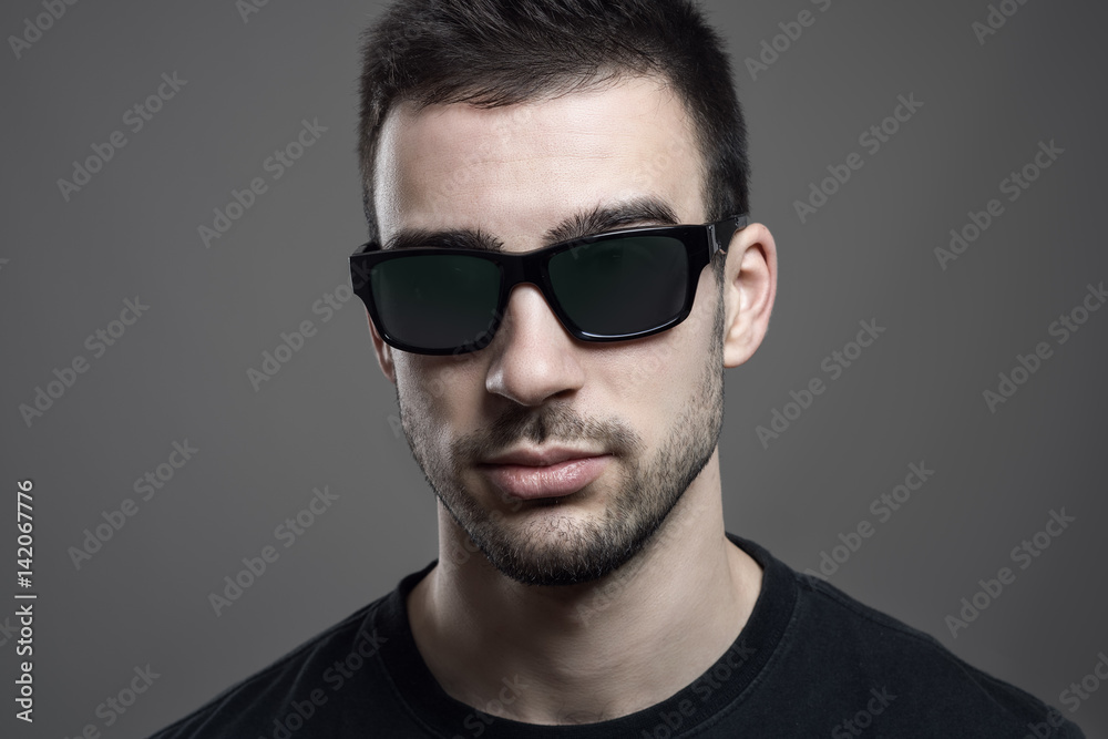 Obraz premium Close up headshot of serious young handsome man wearing sunglasses. Atmospheric contrasty portrait over gray studio background.