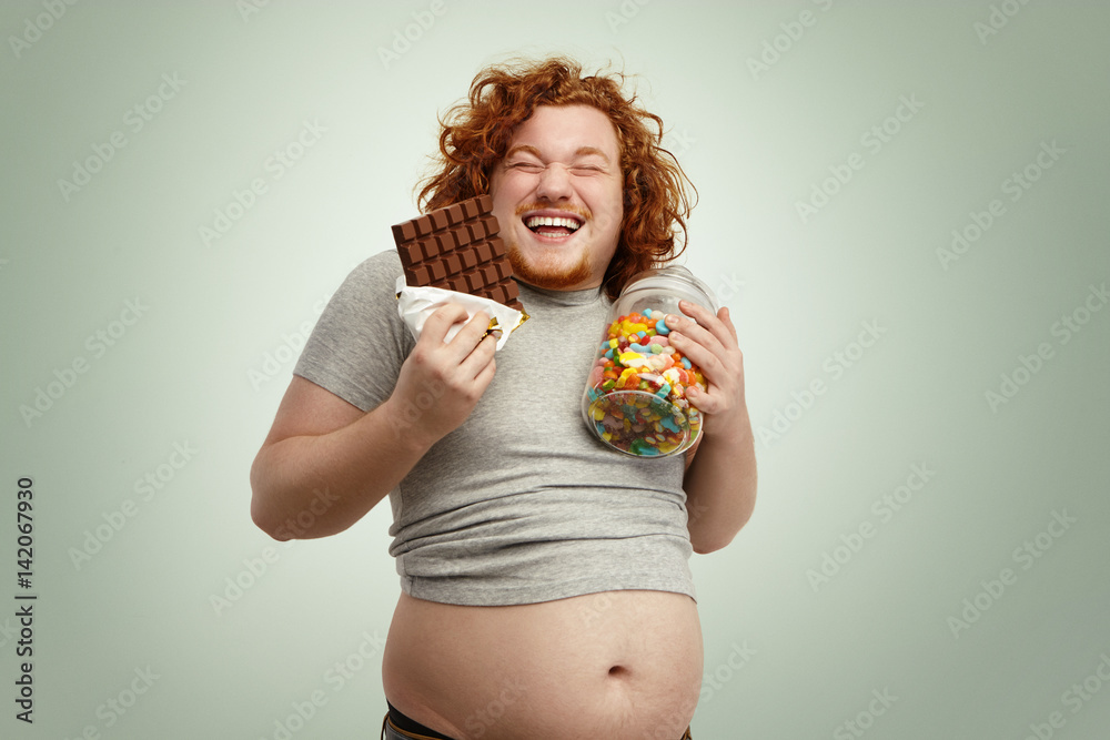 Stockfoto med beskrivningen Indoor shot of fat redhead male holding glass  of sweets in one hand and bar of chocolate in other, laughing, having sly  look, anticipating eating sweets, his belly sticking