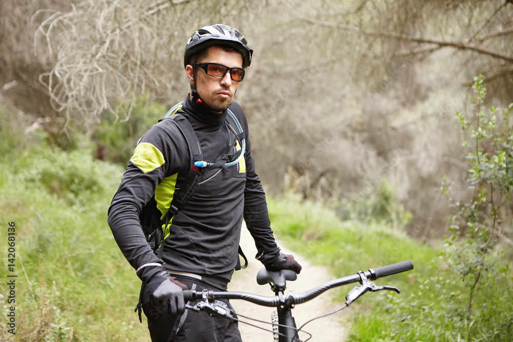 Attractive stylish male biker wearing black cycling clothing, glasses and helmet standing on trail in rural area, holding hand on handlebar of his motor-powered electric bicycle, looking at camera Attractive stylish male biker wearing black cycling clothing, glasses and helmet standing on trail in rural area, holding hand on handlebar of his motor-powered electric bicycle, looking at camera