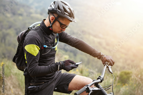 Side view of young European rider standing on top of hill, holding mobile phone, using online application for searching GPS coordinates while riding mootor-powered electric bike in forest on sunny day
