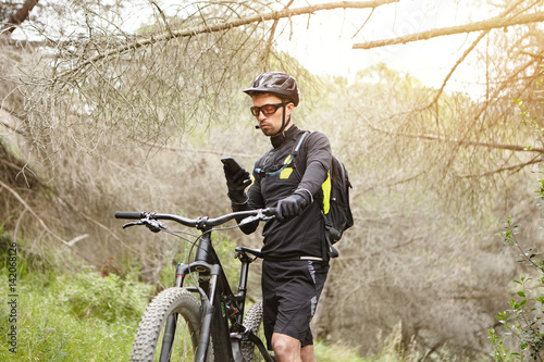 Serious concentrated young cyclist in black clothing studying map on smart phone, holding hand on handlebar of his electric bicycle, searching GPS coordinates to find right way while cycling in woods