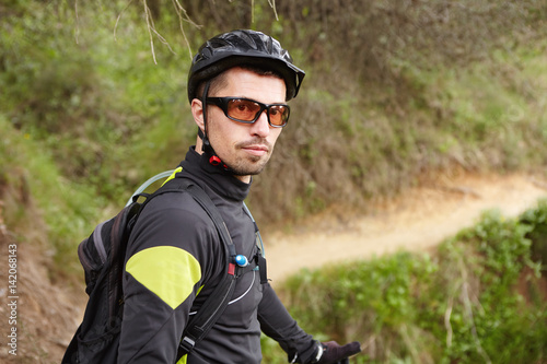 Portrait of handsome confident professional cyclist in sportswear, glasses, helmet and backpack looking at camera, standing on trail in forest, ready for workout. Sports, active lifestyle and extreme
