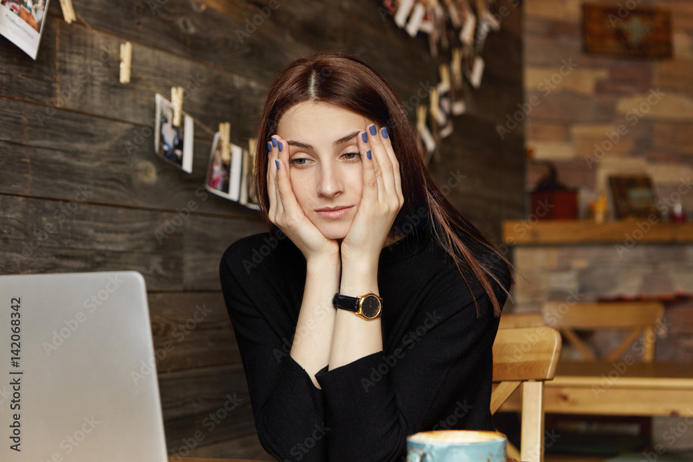 Stressed young Caucasian female freelancer resting face on her hands looking at laptop screen in front of her with bored expression, feeling tired while working remotely at cafe. People and lifestyle Stressed young Caucasian female freelancer resting face on her hands looking at laptop screen in front of her with bored expression, feeling tired while working remotely at cafe. People and lifestyle