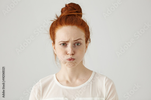 Headshot of attractive funny young female with ginger hair dressed in white blouse feeling displeased or uncomfortable with something, blowing cheeks and frowning. Human face expressions and emotions photo