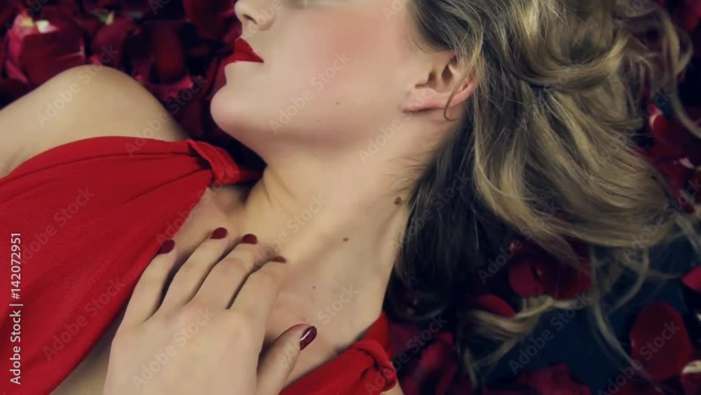 Video footage of  girl in red dress with a beautiful hairdo, lies in petals of red roses, turns slowly her head from the side, looks into the camera smiling, hands touching her body. Valentine's Day.