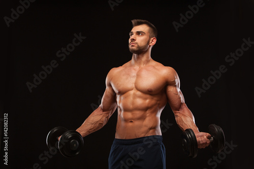 Handsome athletic man in gym is pumping up muscles with dumbbells in a gym. Fitness muscular body isolated on dark background.