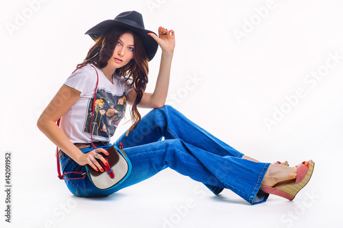 Beautiful young brunette in jeans bell bottom white t-shirt and black hat ,sitting on the floor in the Studio with a small beautiful bag, isolated on white background. photo