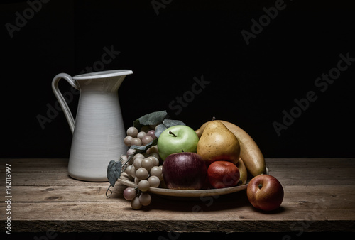 still life of various kinds of fruit and jug of water, with pictorial light on rustic wooden tables on a black background