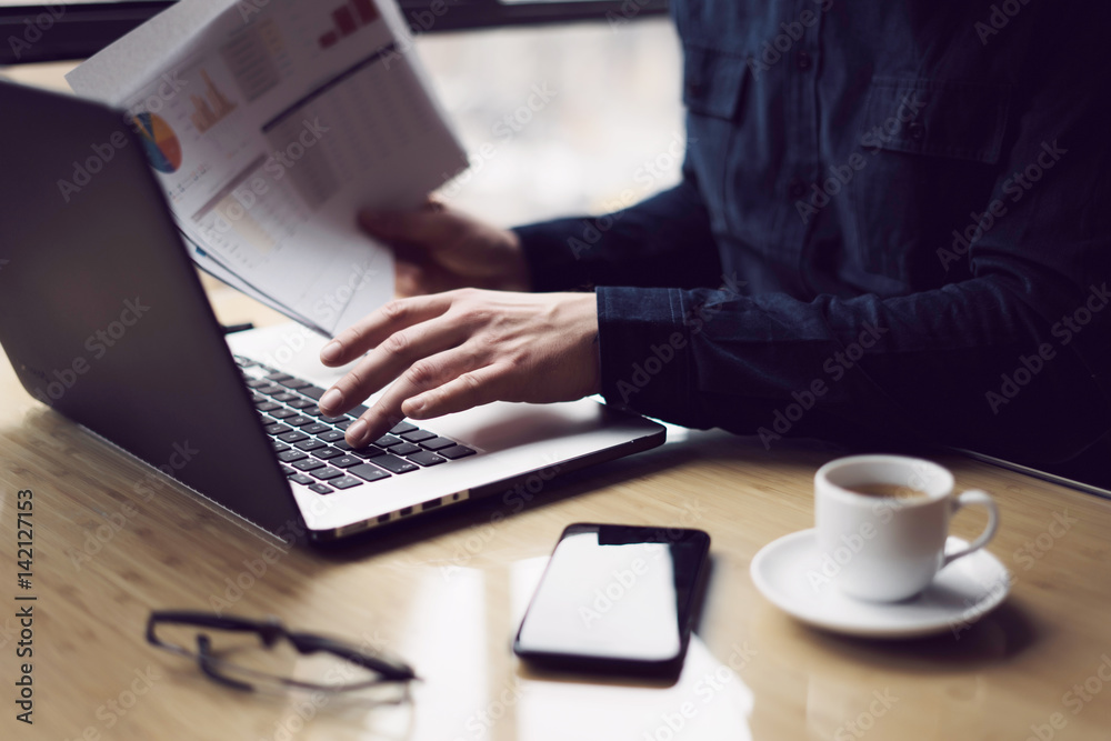 Elegant businessman working at sunny office on laptop while sitting at the wooden table.Man holding paper reports in hands.Blurred background. Elegant businessman working at sunny office on laptop while sitting at the wooden table.Man holding paper reports in hands.Blurred background.