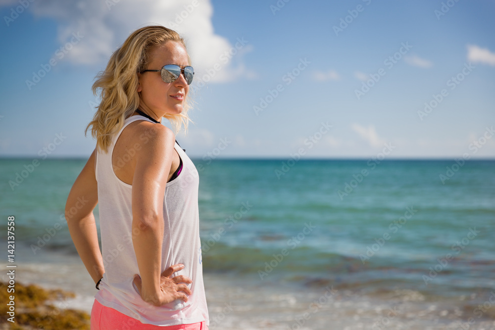 Young sportive woman in sun glasses standing on the beach and looking into distance. Vacations by the sea. Summer camp. Girl relaxing after work out outdoors.
