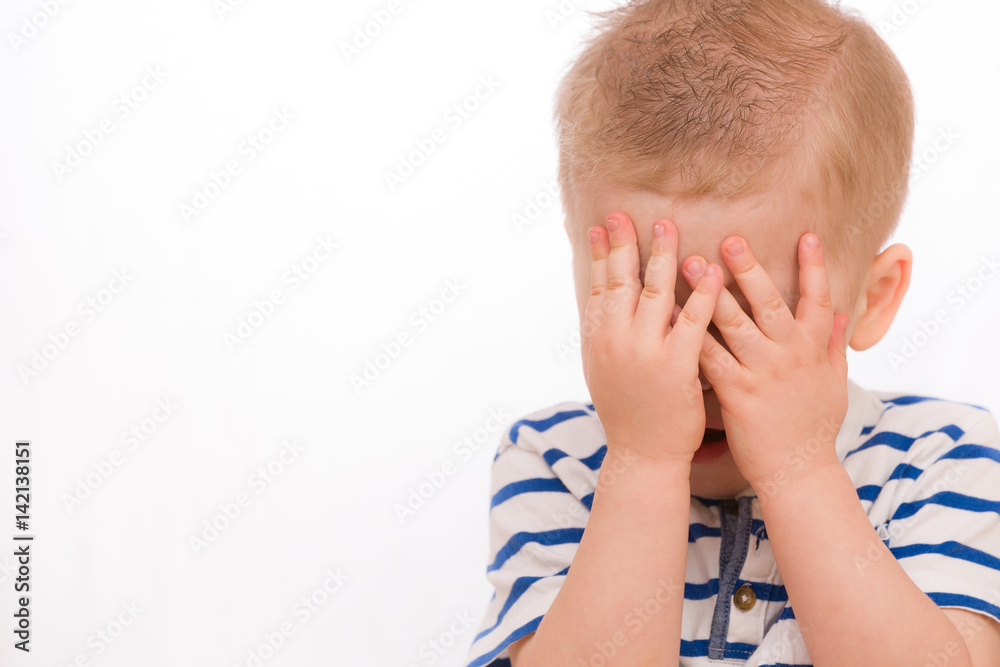 Portrait of a small toddler blond boy in a striped shirt hiding himself with hands. Child crying. Emotions. Portrait of a small toddler blond boy in a striped shirt hiding himself with hands. Child crying. Emotions.