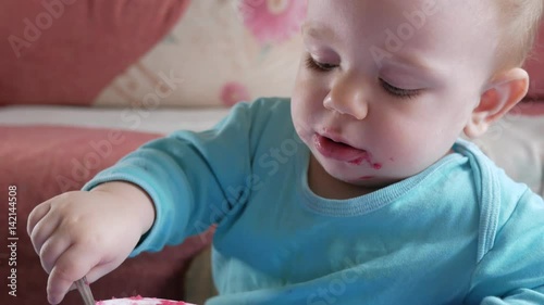 An attractive boy 2 years old eats a red beet salad. The face is smeared with porridge. Sits on the table.
