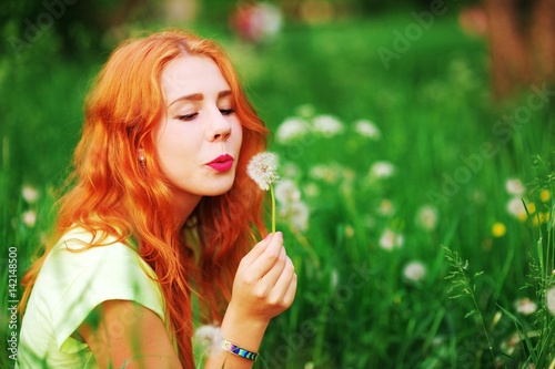 Portrait of young pretty happy red-haired girl sitting on the green lawn and blowing on a dandelion enjoying summer sun