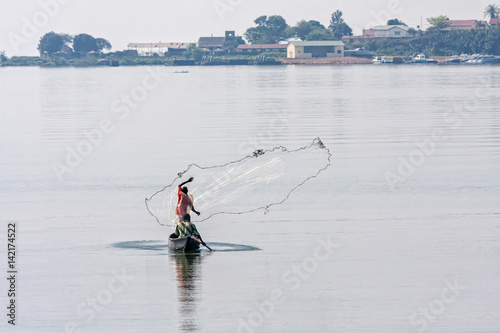 Fisherman in boat casts a net in Victoria Lake bay against hazy bank background. Entebbe, Uganda, Eastern Africa.
