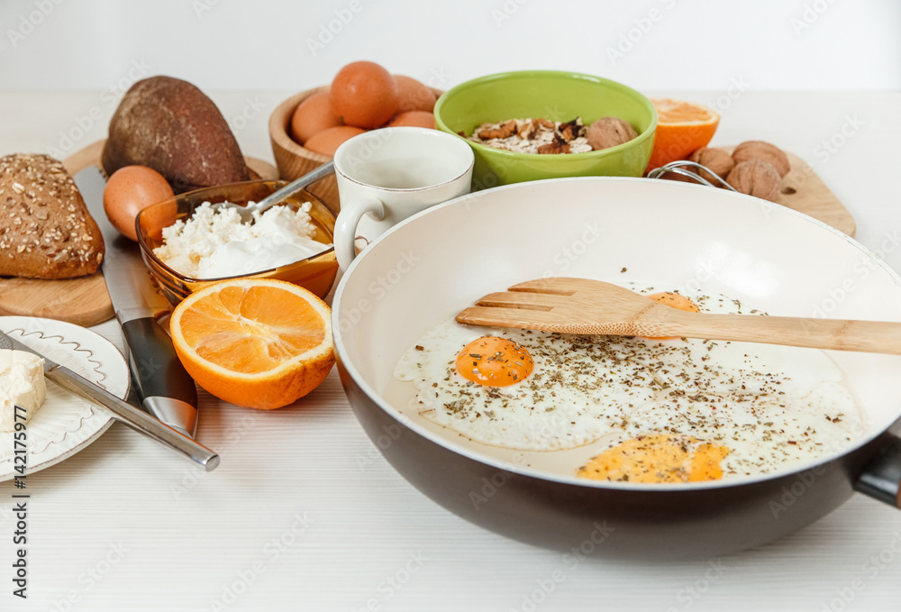 Fried Eggs in the Frying Pan,Breakfast Ingredients.Orange,Bread,Butter.Kitchen Accessories.Cooking Morning Food.White Table.Selective Focus Fried Eggs in the Frying Pan,Breakfast Ingredients.Orange,Bread,Butter.Kitchen Accessories.Cooking Morning Food.White Table.Selective Focus