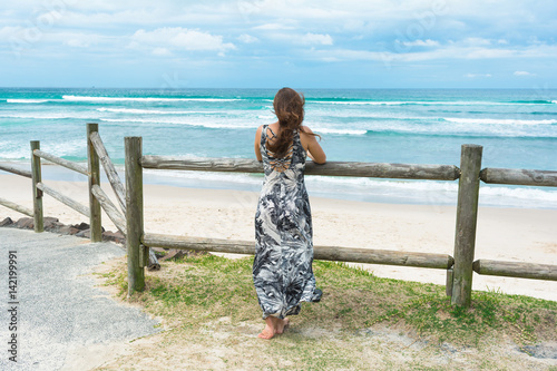 The woman with long dark hair wearing long romantic dress facing on the ocean is looking into the distance on the stormy ocean in Byron Bay, Australia