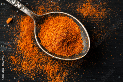 Paprika powder in a metal spoon on a black table, top view, flat lay