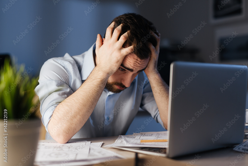 Portrait of exhausted man working overtime with documentation and laptop alone in dark office late at night, clasping head with hands looking stressed Portrait of exhausted man working overtime with documentation and laptop alone in dark office late at night, clasping head with hands looking stressed