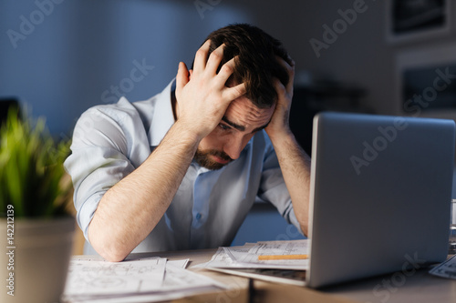 Portrait of exhausted man working overtime with documentation and laptop alone in dark office late at night, clasping head with hands looking stressed