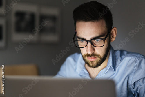 Portrait of handsome bearded man wearing glasses working with laptop in dark room late at night, his face lit up by screen photo