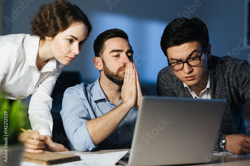 Portrait of three colleagues, one of them Asian, working overtime in dark office late at night: collaborating on project and using laptop trying to finish by deadline