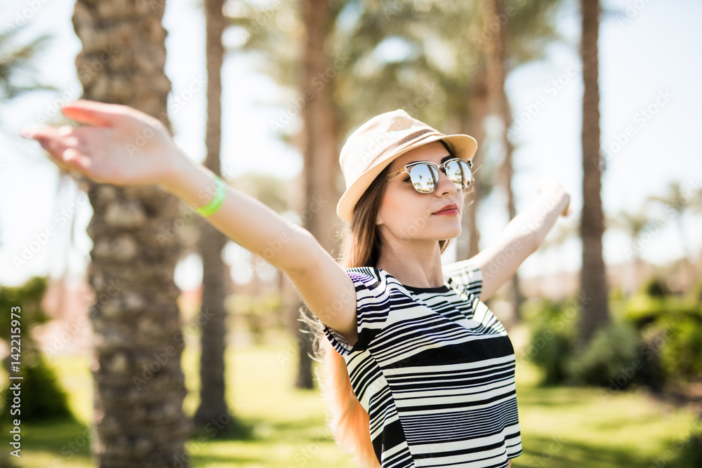 Smiling relaxed young woman standing with raised hands in hat and sunglasses on summer resort