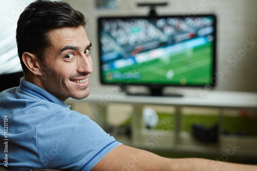 Portrait of handsome man smiling, turning to look at camera while watching football match at home