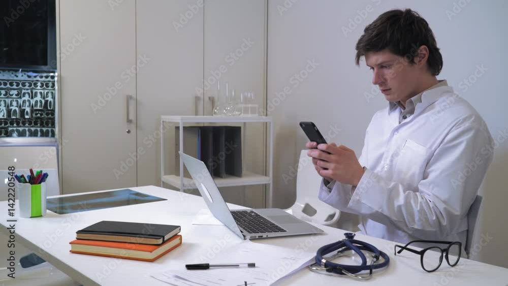 Doctor using smartphone in office. Caucasian young man wearing in white coat sitting at the working place messaging on smartphone.