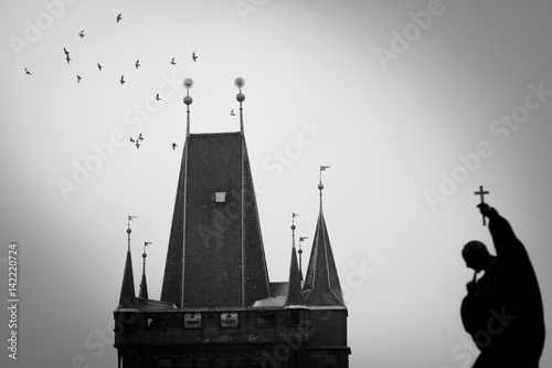Photography Shadow of sculpture with cross in hand and flying birds around the tower of Charles Bridge