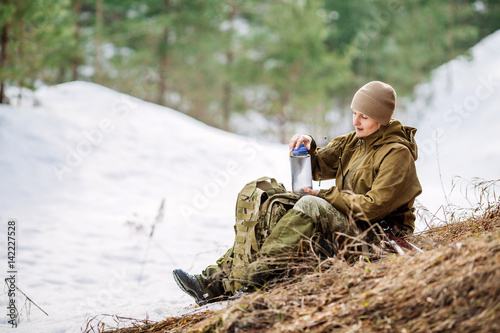 Hunter girl sitting with backpack and drinking water outdoor snow forest in background.