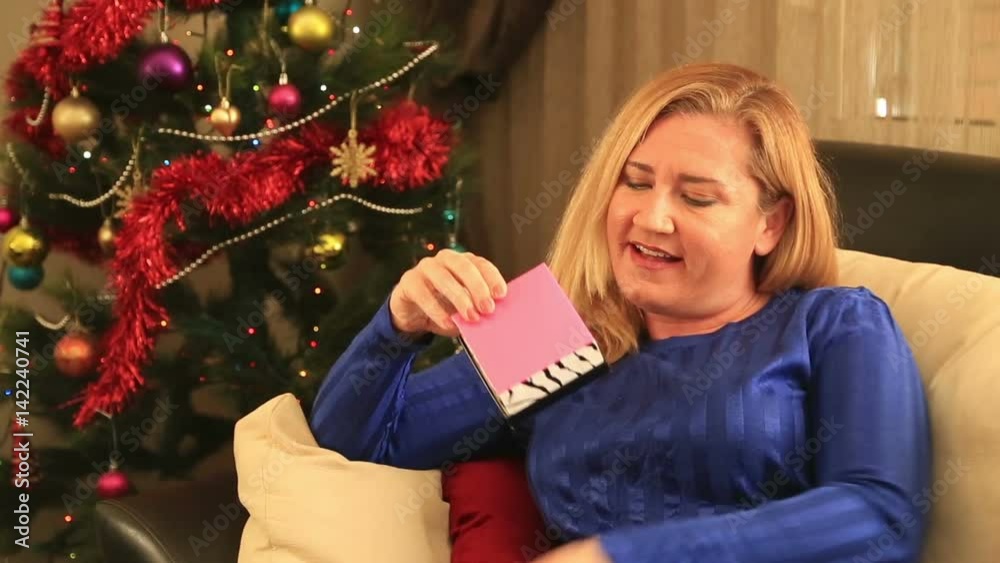 Attractive woman opening christmas present disappointed and unhappy sitting on a sofa front of a christmas tree Attractive woman opening christmas present disappointed and unhappy sitting on a sofa front of a christmas tree