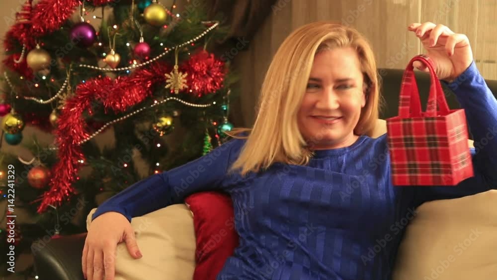 Smiling , happy blond woman displaying a red Christmas gift in her hands in front of a christmas tree. Christmas, x-mas, winter, happiness concept Smiling , happy blond woman displaying a red Christmas gift in her hands in front of a christmas tree. Christmas, x-mas, winter, happiness concept