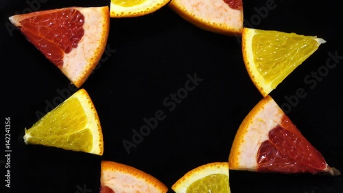 Excellent top view of orange and red grape-fruit sector slices, put in sun on black dish, rotating clockwise. Concentric flat lay of yummy pieces of dessert on dark background with amazing texture